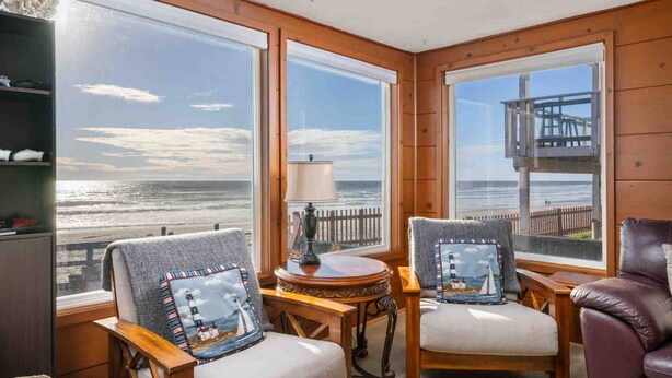 Een kamer met twee stoelen gericht op een groot raam met uitzicht op het strand, een bijzettafeltje met een lamp, en een houten terras buiten.