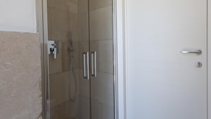 Une douche fermée par des parois de verre, dotée d’un panneau de commande, une porte blanche avec une poignée et un mur recouvert de carreaux.