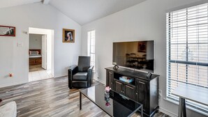 Een ruime woonkamer met houten vloeren, een glazen salontafel, een zwarte leren fauteuil, een donkere houten entertainmentkast en een groot raam met luxaflex.