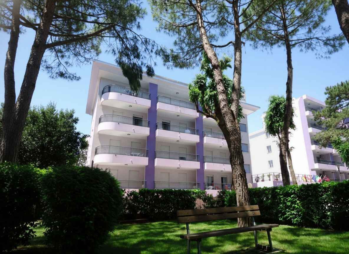 A multi-story building with balconies, surrounded by trees and a grassy area with a bench.