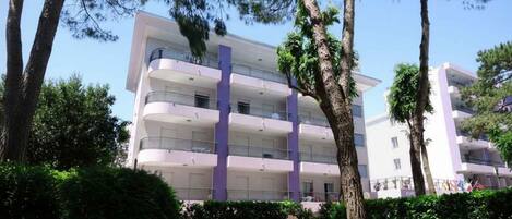A multi-story building with balconies, surrounded by trees and a grassy area with a bench.
