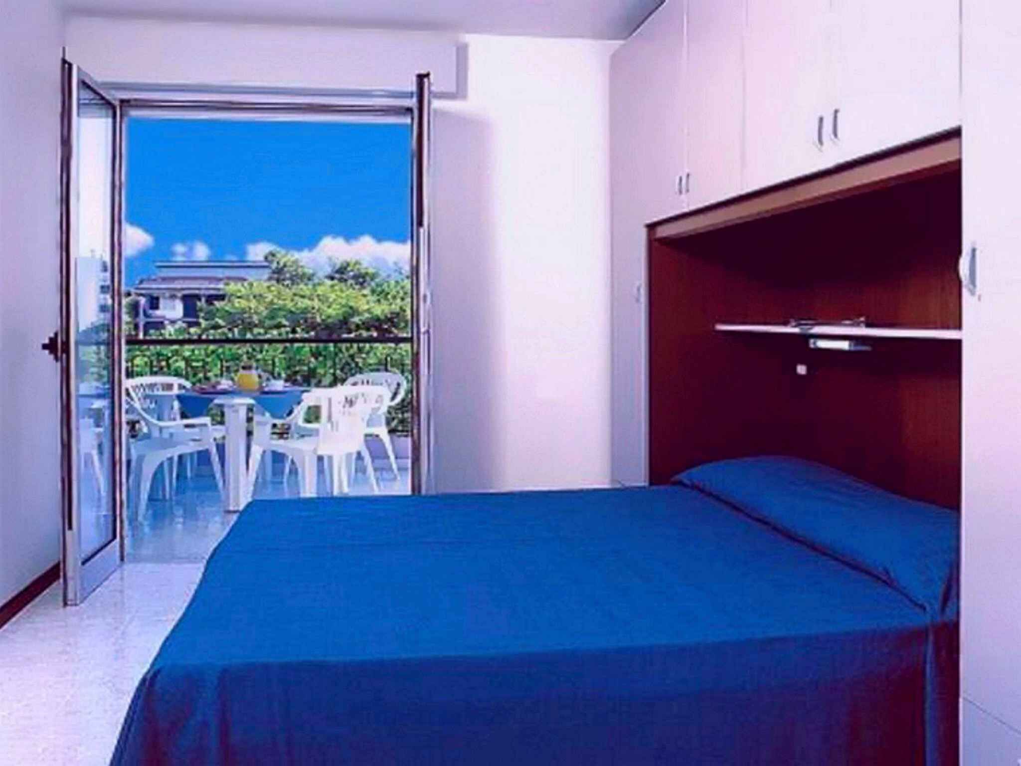 Ein Zimmer mit Bett, einem Balkon mit Tisch und Stühlen sowie Blick auf Gebäude und Grünflächen.