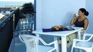 A balcony with a white table and chairs, a person sitting, and a view of the city. - Studio with air conditioning, balcony and pool (Bibione)
