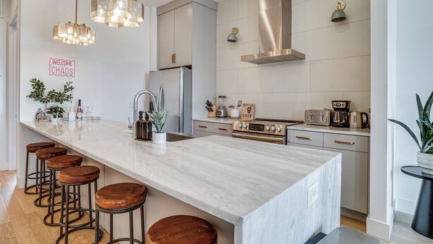 Una cocina moderna con una isla grande, taburetes de bar, electrodomésticos de acero inoxidable e iluminación colgante.