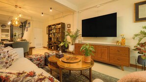 Een woonkamer met een bank, salontafel en televisie. Er staat een boekenkast en een bloempot.
