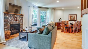 A living room with a stone fireplace, a sofa, a dining table with chairs, and a large window with curtains. - Game Room & Hot Tub! Dog-Friendly Arnold Retreat (Arnold)