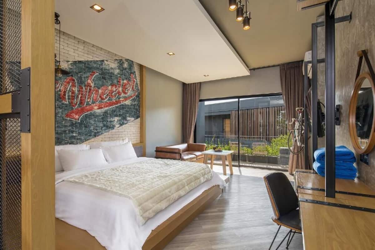 Una habitación de hotel moderna con una cama grande, un área de descanso con una silla y una mesita, un balcón con vistas y una pared con un cartel de 'Wheelers'.