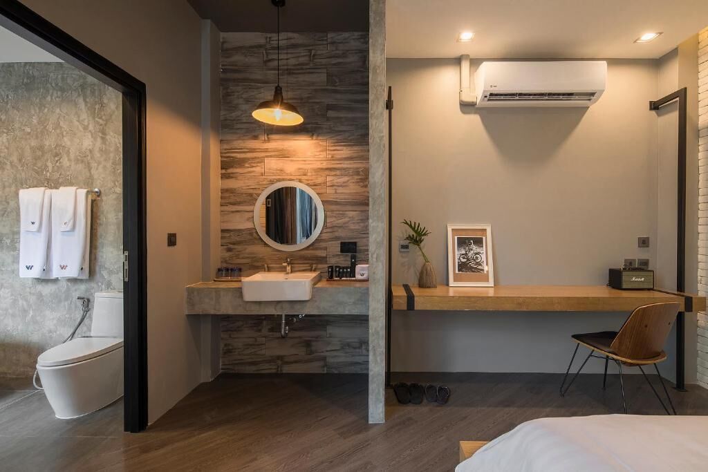 Uma casa de banho moderna com parede de pedra, um espelho redondo, uma pia, um sanitário e uma cadeira.