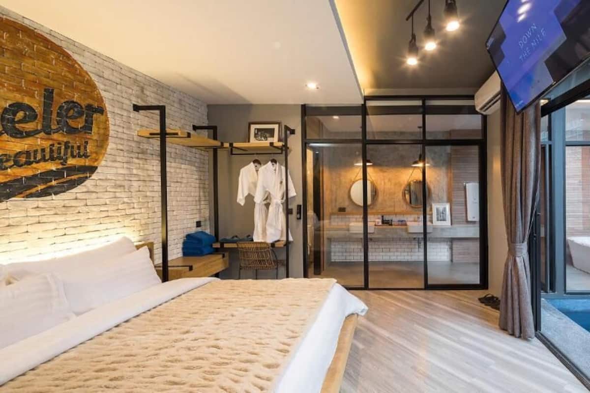 Una habitación de hotel moderna con una cama grande, una pared de ladrillo, una puerta de vidrio que conduce a un baño y un televisor montado en la pared.