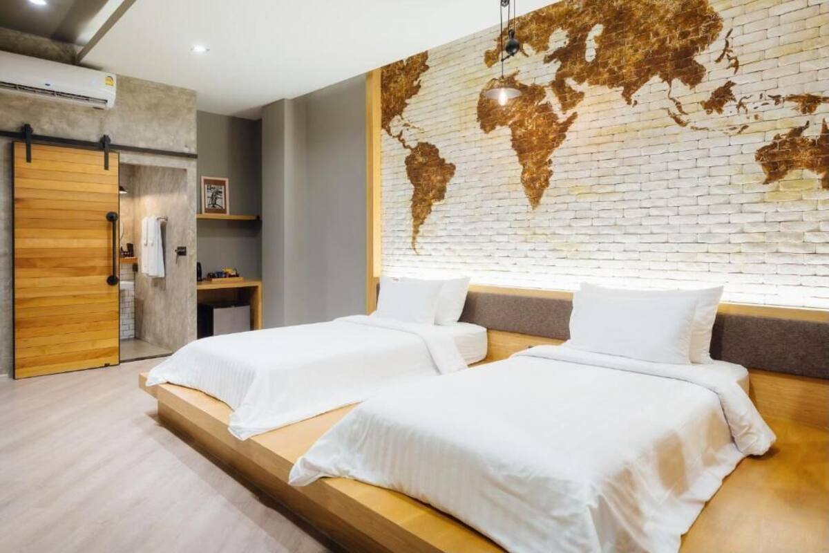 Habitación de hotel con dos camas, un mural de un mapa mundial, una puerta de madera y un estante pequeño con artículos de tocador.