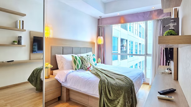 A modern hotel room with a bed, bedside tables, a TV, and a large window with a city view. - Casa By SanLiving - 2BR - Kasablanka (Jakarta)