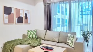 A modern living room with a sectional sofa, a glass coffee table, and a large window with curtains. - Casa By SanLiving - 2BR - Kasablanka (Jakarta)