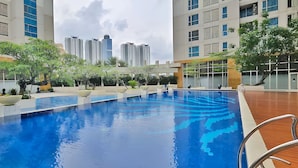 A swimming pool with a wooden deck, surrounded by greenery and modern buildings in the background. - Casa By SanLiving - 2BR - Kasablanka (Jakarta)
