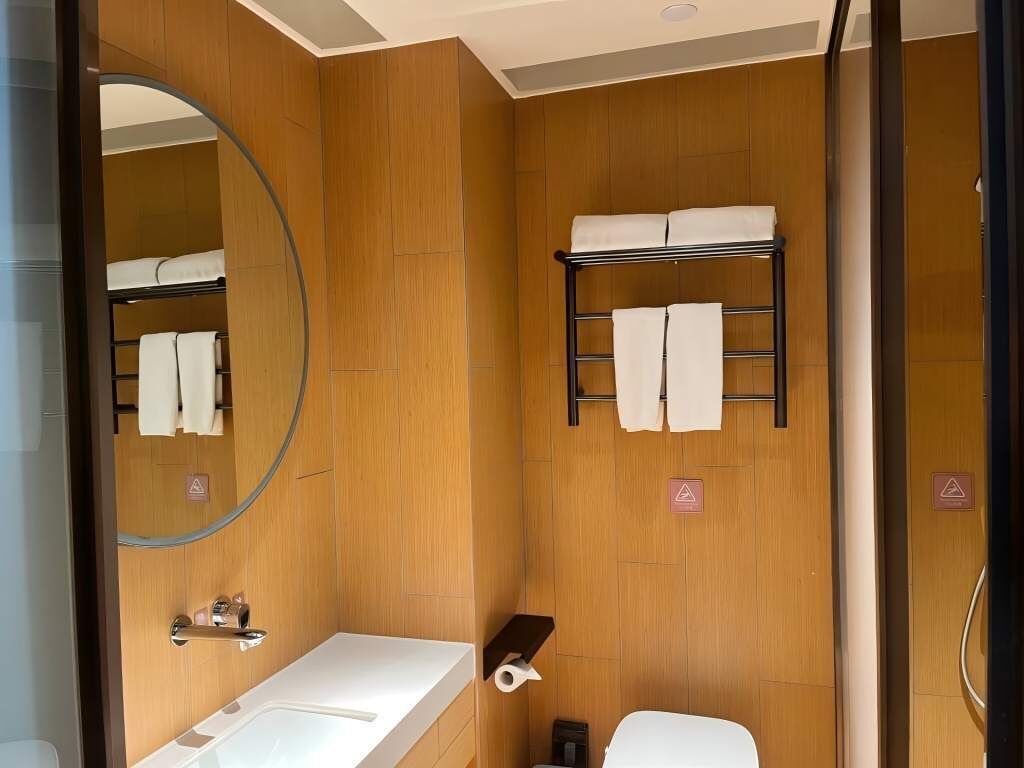 A compact bathroom with a wooden wall, a large mirror, a sink, a toilet, and a towel rack.