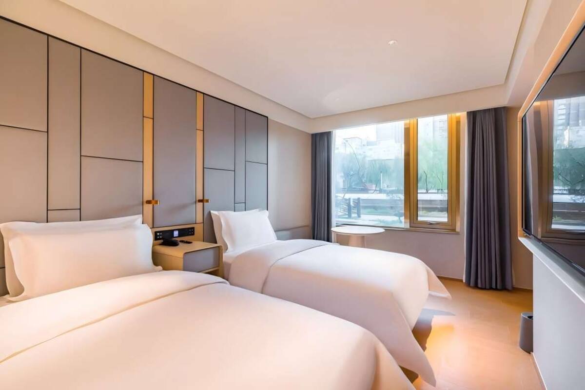 Habitación de hotel moderna con dos camas, una mesita de noche, una pequeña mesa redonda y un ventanal grande con cortinas.