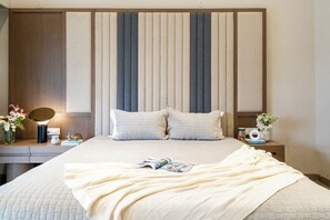 A neatly made bed with a quilted cover, a bedside table with a lamp, a vase with flowers, and a wall-mounted shelf with decorative items. - Staymaster Casa Nivara (Siolim)