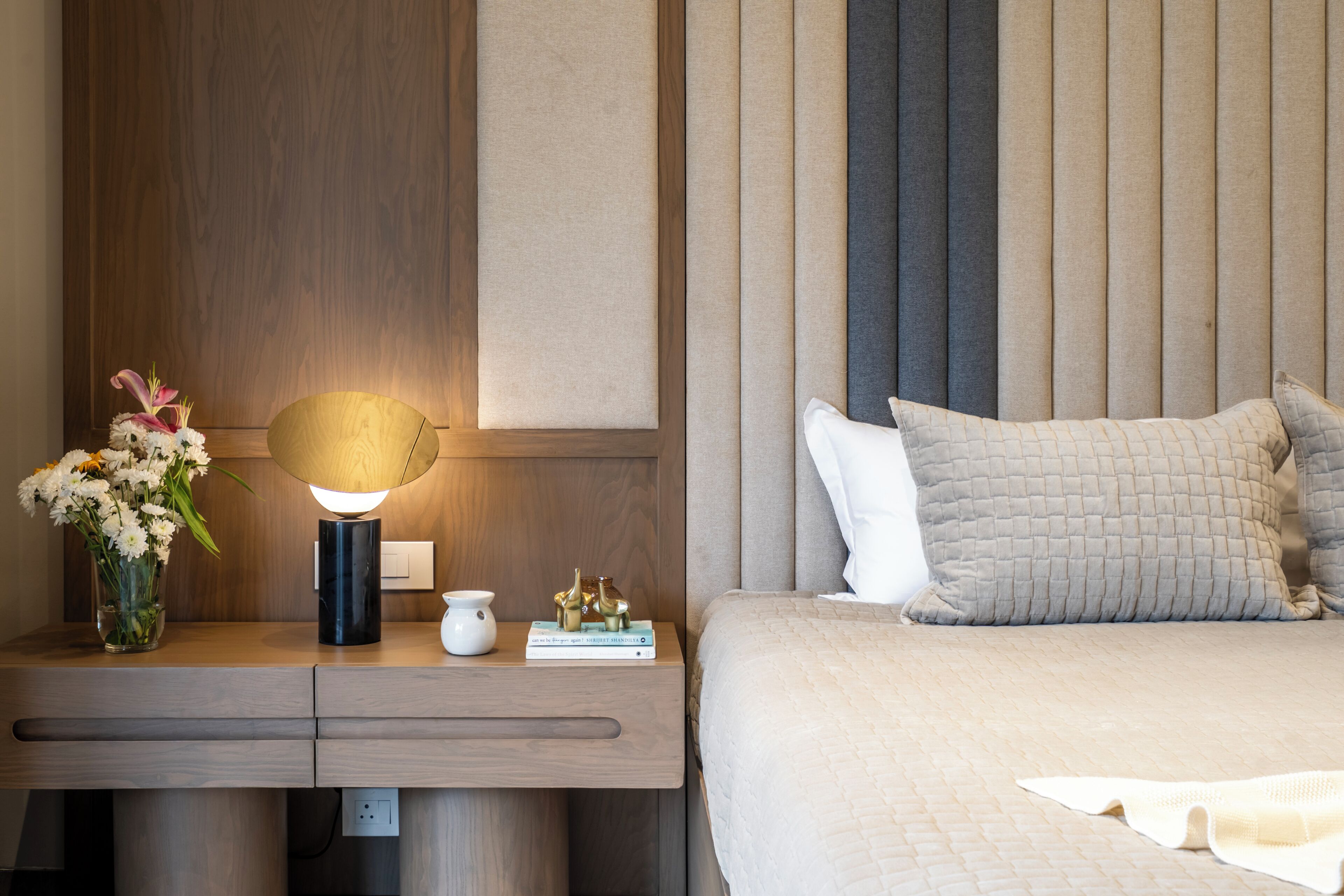 A neatly arranged bedroom with a wooden nightstand, a bedside table, a lamp, a vase with flowers, a book, and a bed with pillows and a quilt.