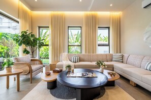 A modern living room with a sofa, coffee table, and chairs, large windows with curtains, and indoor plants. - Staymaster Casa Nivara (Siolim)