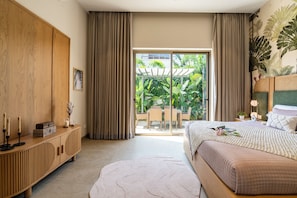A bedroom with a bed, a wooden wardrobe, a bedside table with candles, and a view of a garden through a glass door. - Staymaster Casa Nivara (Siolim)