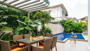 A patio area with a dining table and chairs, a pool, and lush greenery. - Staymaster Casa Nivara (Siolim)