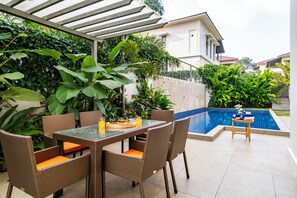 Un patio con mesa y sillas, piscina y abundante vegetación.