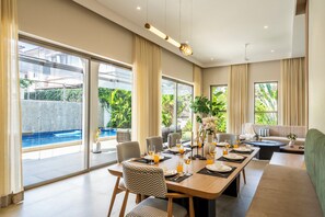 A modern dining area with a pool view, large windows, and a cozy seating area. - Staymaster Casa Nivara (Siolim)