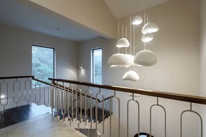 A staircase with a wooden handrail and metal balusters, leading to a landing with large windows and hanging pendant lights. - Staymaster Casa Nivara (Siolim)
