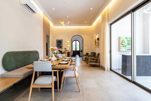 A modern dining area with a long table, chairs, and a sofa. There is a large window with curtains, a ceiling fan, and an air conditioning unit. - Staymaster Casa Nivara (Siolim)