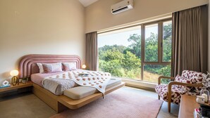 A bedroom with a bed, bedside table, lamp, and a view of greenery through the window. - Staymaster Casa Nivara (Siolim)