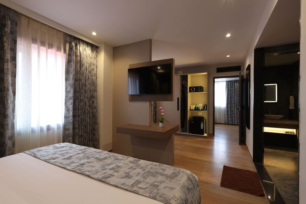 A hotel room with a bed, a TV mounted on the wall, a small desk with a plant, and a bathroom visible through an open door.