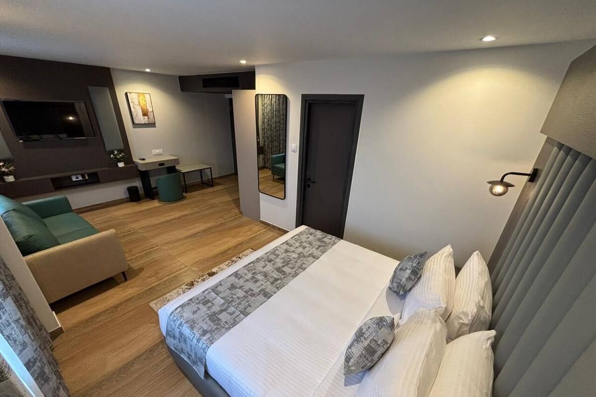 A modern hotel room with a large bed, a seating area with a sofa and armchair, a small desk, and a mirror.