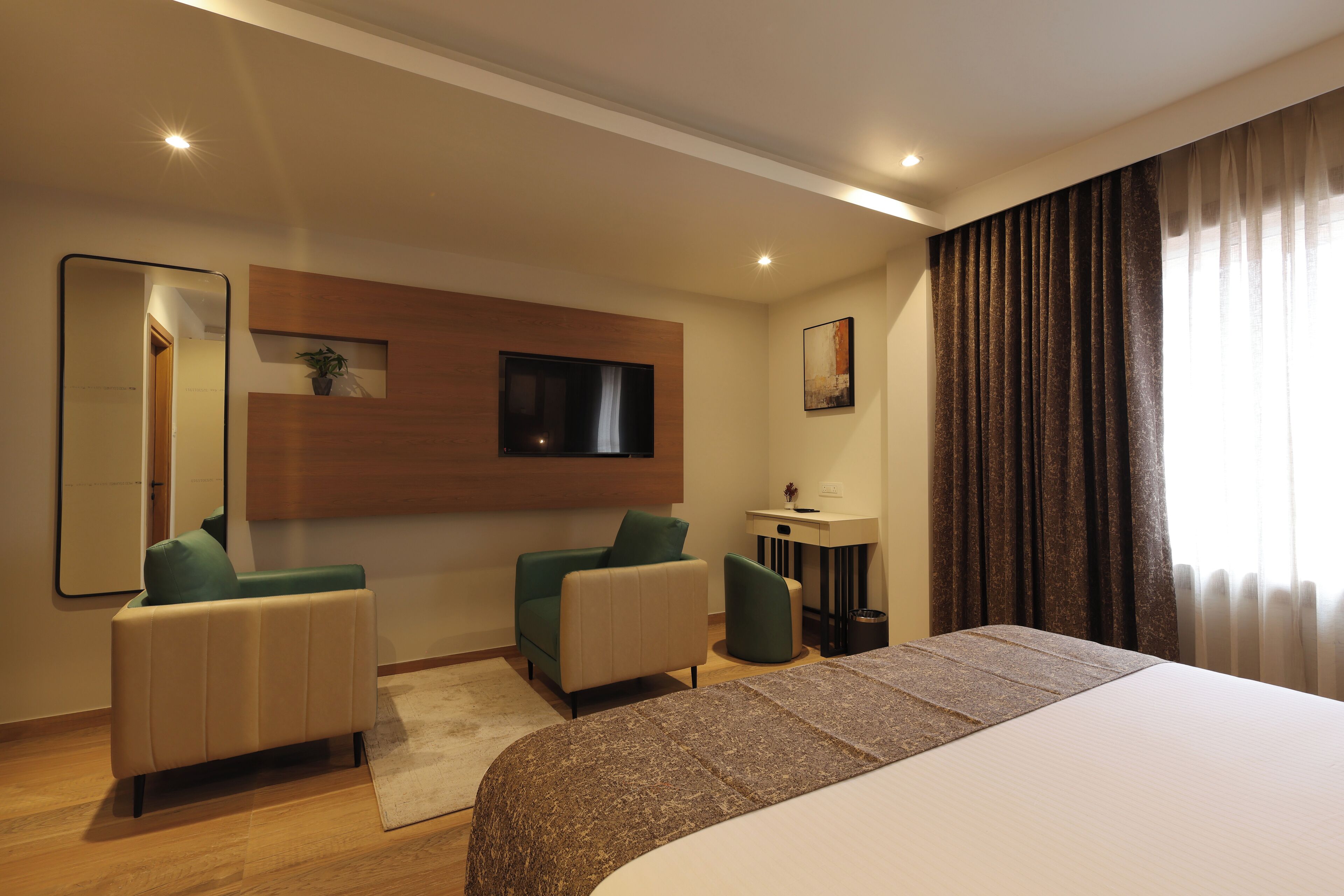 A hotel room with a large bed, two armchairs, a TV mounted on the wall, a small table with a plant, and a window with curtains.