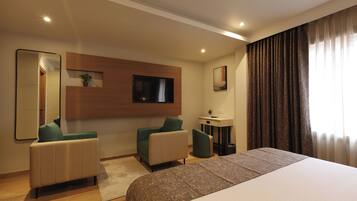 A hotel room with a large bed, two armchairs, a TV mounted on the wall, a small table with a plant, and a window with curtains.
