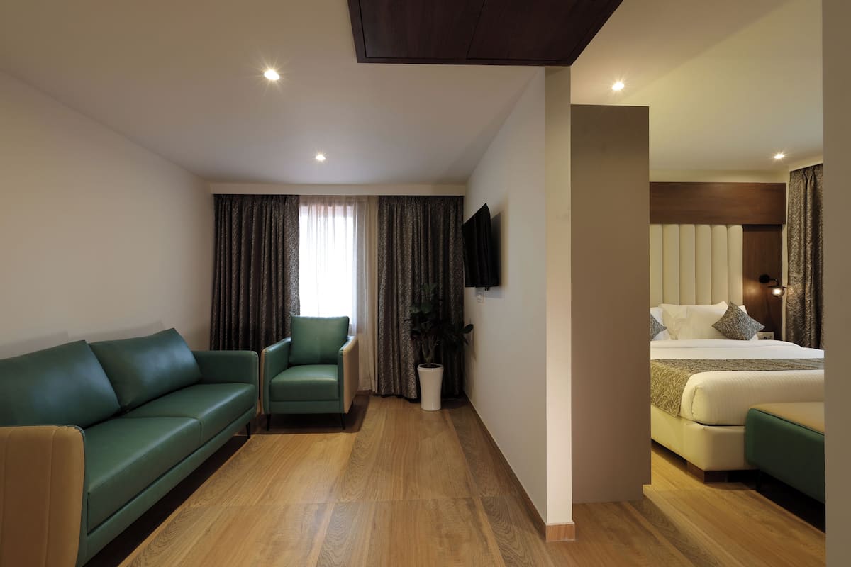 A hotel room with a green sofa, a green armchair, a bed with white bedding, a potted plant, and a flat-screen TV mounted on the wall.