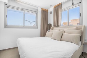 A bedroom with a large bed, two windows with curtains, a hanging light fixture, and a view of a building outside. - Sunlit Desert View Modern Retreat (Eilat)