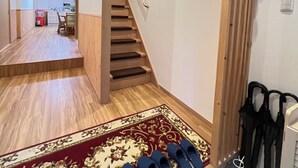 Un pasillo con piso de madera que lleva a una escalera, una alfombra con diseño rojo y dorado, y una fila de pantuflas azules.