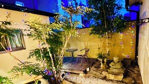 Uma trilha de pedras leva a uma área de estar com mesa e cadeiras, cercada por plantas em vasos e luzes decorativas.