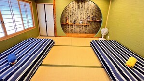 Um quarto com tatames, dois futons de tatame, teto de madeira, porta deslizante e prateleira com objetos decorativos.