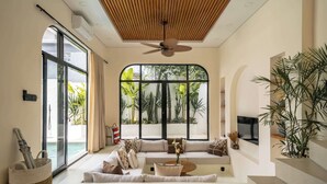 Ein modernes Wohnzimmer mit einem großen Sofa, einem Couchtisch und einem Deckenventilator. Ein großes Fenster bietet Blick auf Grünflächen und einen Pool.