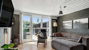 Un salon moderne avec un canapé gris, un téléviseur et une grande fenêtre donnant sur un paysage verdoyant.