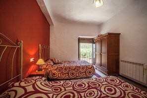 Ein Schlafzimmer mit Bett, Nachttisch, Lampe, Kleiderschrank und einem Fenster mit Blick ins Grüne.