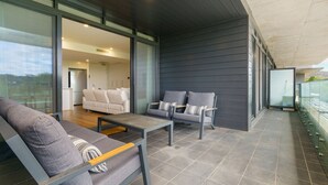 A modern living room with a sofa, two armchairs, and a coffee table. There is a glass door leading to another room with a sofa and a television. The room has a dark wooden floor and a large window. - Premium Apartment with Scenic Views near City (Campbell)