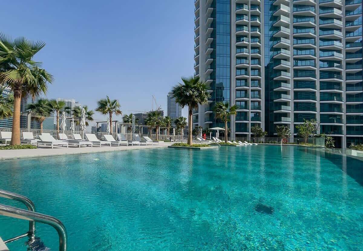 Una piscina con sillones de descanso, palmeras y un edificio alto al fondo.
