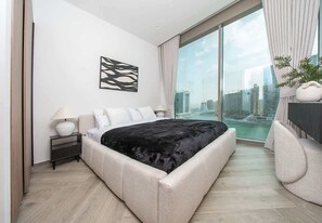 A modern bedroom with a large bed, a nightstand, a lamp, and a view of a cityscape through large windows. - Canal Views Apt W Balcony Pool & Gym Access (Dubai)