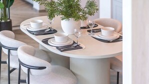 Une table à manger dressée avec de l’assiette, des bols et des couverts blancs, entourée de chaises à la structure noire et au revêtement en tissu clair.