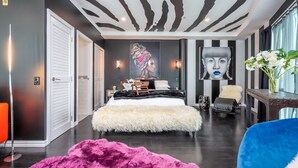 Un dormitorio moderno con techo a rayas de cebra, una cama grande con cubierta blanca esponjosa, una silla azul y una alfombra rosa mullida.