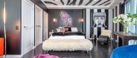 A modern bedroom with a zebra-striped ceiling, a large bed with a fluffy white cover, a blue chair, and a pink furry rug.