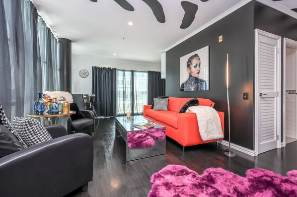 Un salon moderne avec un canapé en cuir noir, un canapé rouge et une table basse en verre. Il y a une grande fenêtre avec des rideaux, une horloge au mur et une porte sur la droite.