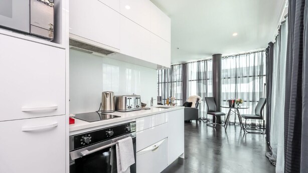 Una cocina moderna con armarios blancos, horno empotrado y cafetera. Hay un comedor con mesa y sillas, y ventanas amplias que ofrecen luz natural.