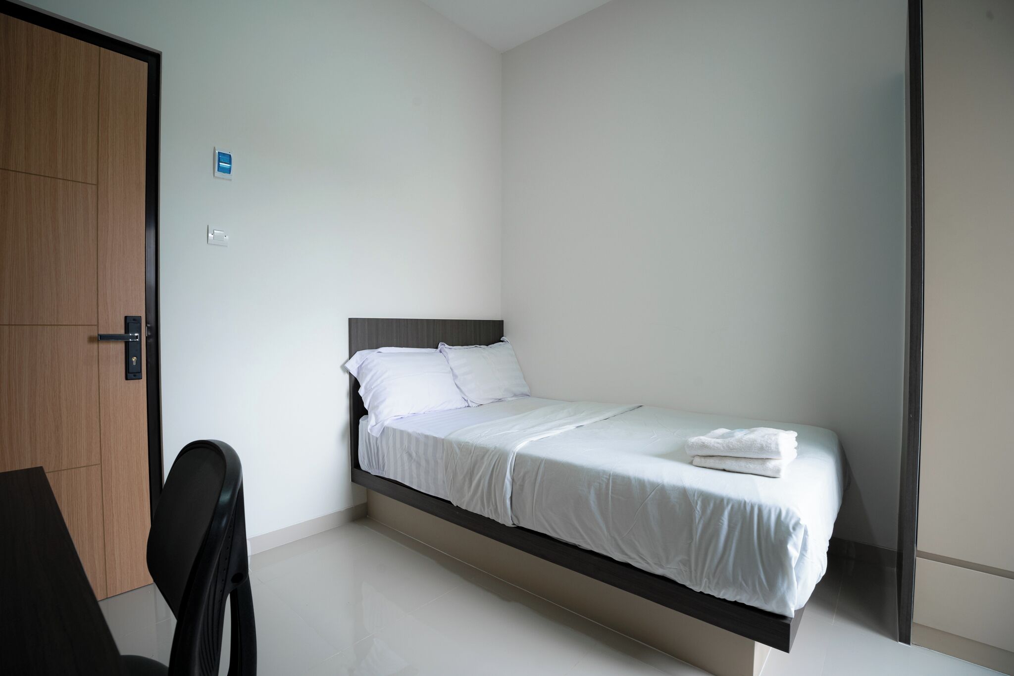 Una cama individual con ropa blanca, una puerta de madera y una silla negra en una habitación minimalista.
